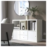 Modern living room with a white ikea KALLAX bookshelf, round mirror, and grey chair. The bookshelf holds books, a lamp, and a vase with greenery.