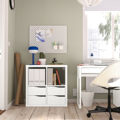 KALLAX shelving with books, plant, lamp, and notes. White desk with computer and chair near window.