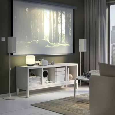 Modern living room with KALLAX shelf, speakers, and forest scene on large screen, grey sofa, coffee table.