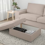 Beige KIVIK footstool with storage, magazines, blanket, next to sofa and side table with vase and teacup. Bright, airy window.