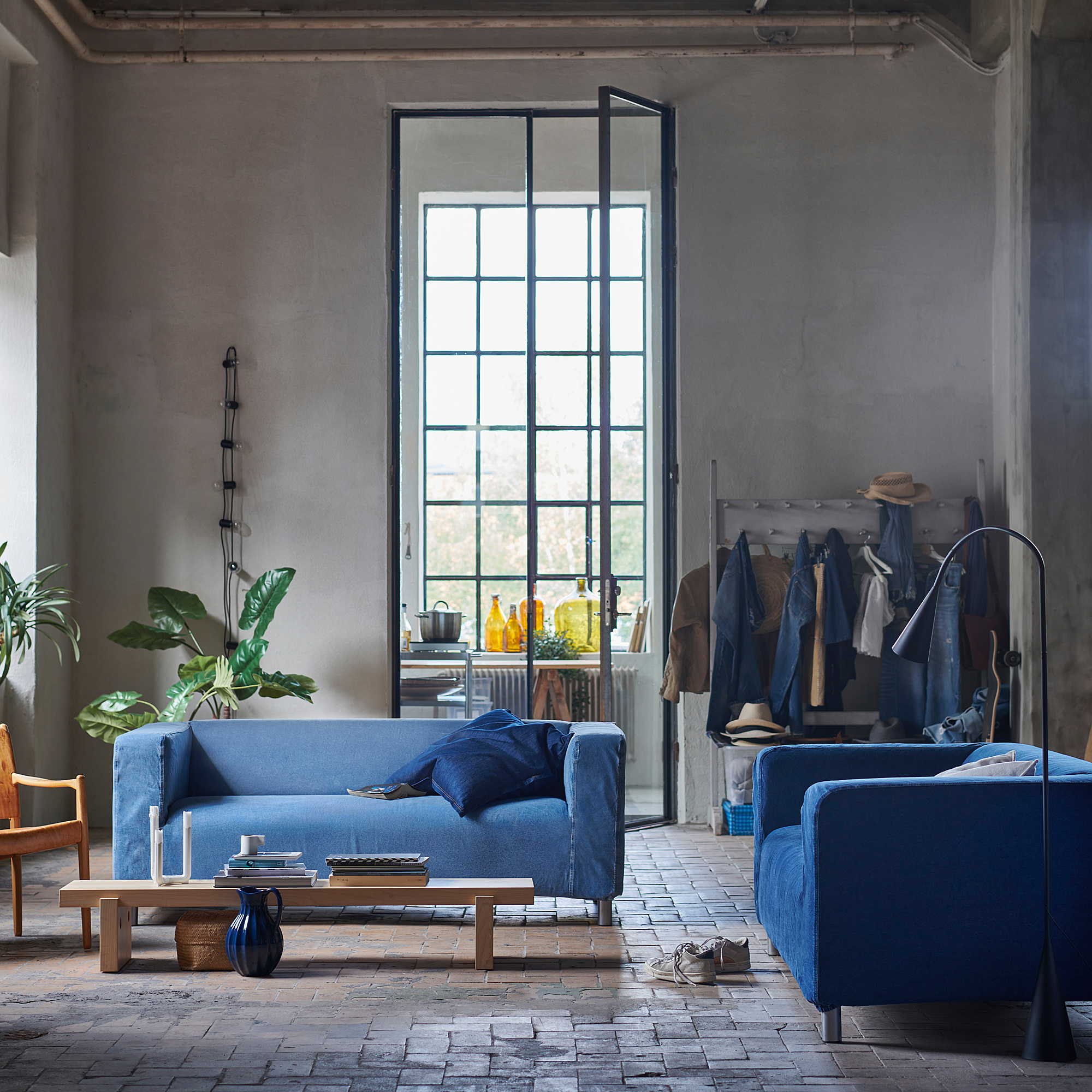 Cozy room with blue KLIPPAN sofas, brick floor, large window, coats, and plants.