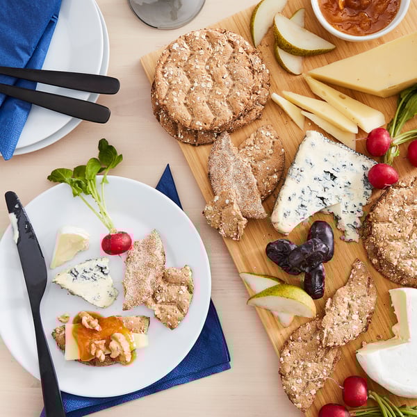A cheese board with KNÄCKEBRÖD FLERKORN crispbread, various cheeses, fruits, and radishes.