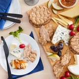 A cheese board with KNÄCKEBRÖD FLERKORN crispbread, various cheeses, fruits, and radishes.