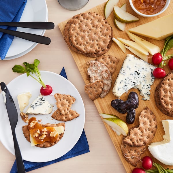 Top view of a snack setup with KNÄCKEBRÖD RÅG crispbread, cheeses, crackers, fruit, and radishes on a wooden board and plate.