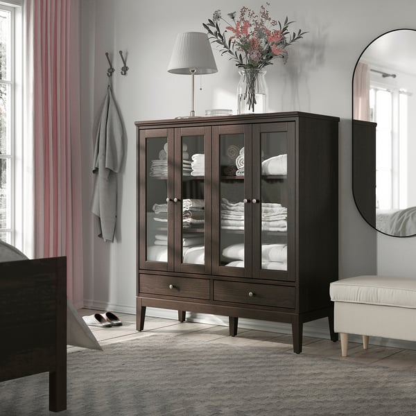 Modern bedroom with KNAPPHIRS dark wood cabinet, large, holding white linens. Topped with lamp and flowers. Mirror and bench beside it.