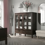Modern bedroom with KNAPPHIRS dark wood cabinet, large, holding white linens. Topped with lamp and flowers. Mirror and bench beside it.