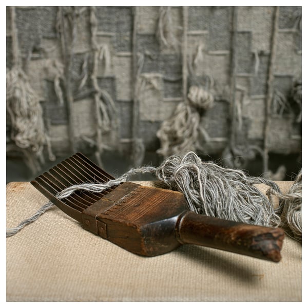 Wooden tool with grey string on beige cloth, background has grey textured wall with hanging strings.