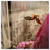 Person weaving rug using loom, holding tools, wearing pink. Traditional craft, handmade.