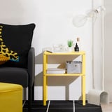 Yellow KNARREVIK side table next to gray chair, holding magazines, adding color to room.