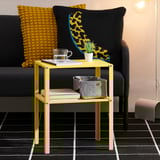 Bright yellow side table with shelves, perfect for holding drinks, plants, and books. Decorated with colorful pillows and a modern lamp.