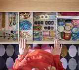 Wooden jewelry drawer by KOMPLEMENT, holding bracelets, necklaces, rings, and accessories. Partially open with hands reaching in.