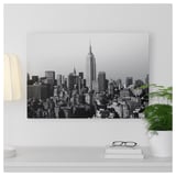 Black and white cityscape photo print on metal, displayed above a white shelf with a plant and books.