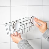 Person holding a silver KROKFJORDEN shower basket with two white soap dishes, against a white tiled wall.