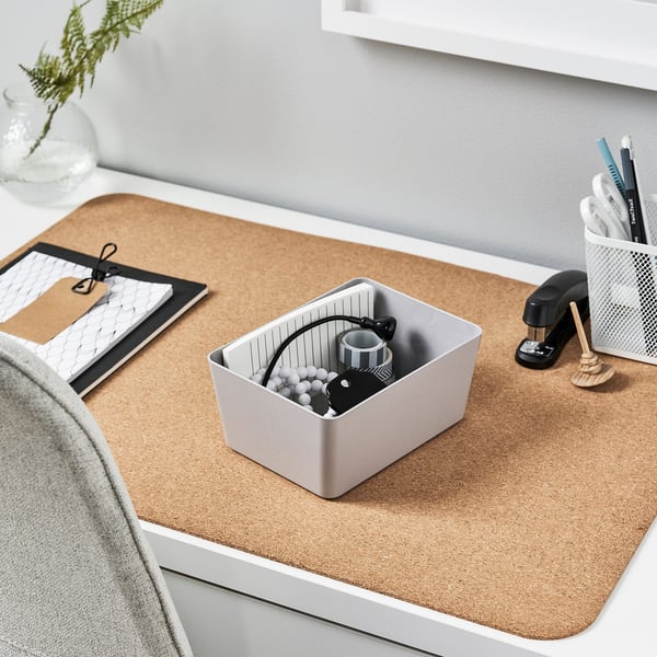 A KUGGIS storage box in grey on a cork desk mat. It holds a mug, keys, and cables. The box is practical and fits neatly with other items on the desk.