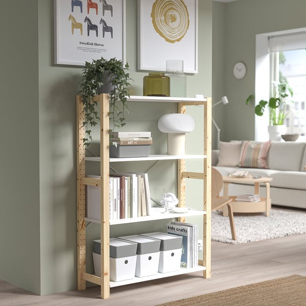 Light wooden bookshelf with white shelves, holding plant, books, and boxes. Situated in a cozy living room with green walls and natural light.