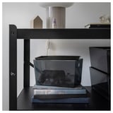 Black KUGGIS storage box with lid on black shelf, holding a black game controller. Modern, sleek design made of recycled pet plastic.