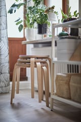 Modern, wooden stools stacked, placed near a window with green plants, brightening cozy space.