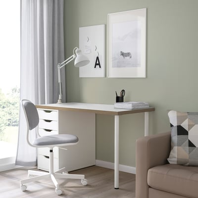 Modern home office with white LAGKAPTEN desk, window view, three drawers, gray chair, lamp, minimalist posters, beige couch, geometric pillow.