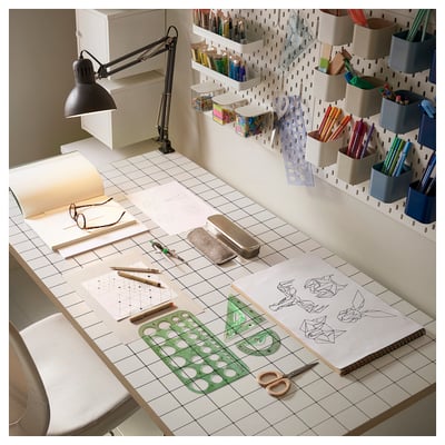 A well-organized crafting desk with white surface, green ruler, black lamp, various stationery items in containers, art supplies, and drawings.