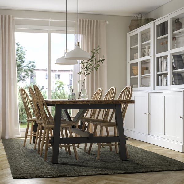 Modern dining room with dark green rug, wooden table, chairs, lamp, bookshelf, and large window.