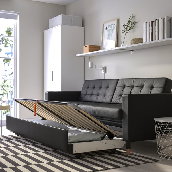 Modern living room with black leather sofa-bed, wooden legs, storage, and oak shelves with books and plants.