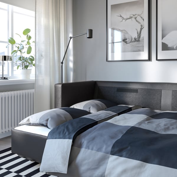 Modern bedroom with grey bed, striped rug, wall art, floor lamp, and potted plant by window.