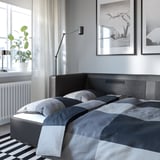 Modern bedroom with grey bed, striped rug, wall art, floor lamp, and potted plant by window.
