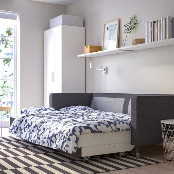 Modern bedroom with landskröna convertible sofa-bed against wall, featuring gray upholstery with patterned bedding, white cabinet, and striped rug.