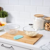 Glass bowls with dough, spatula, flour, plant on counter.