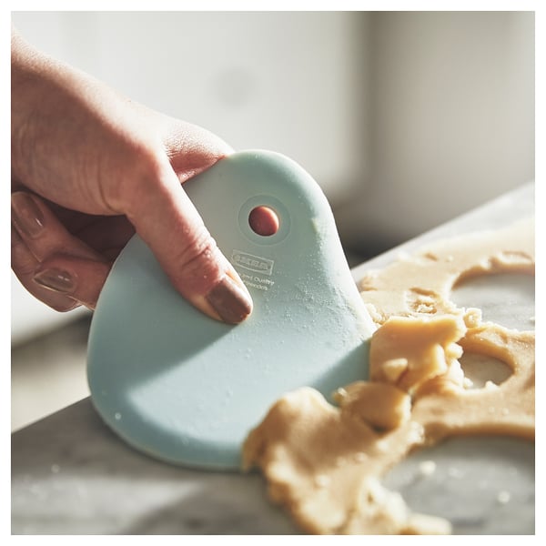 A hand uses a light blue LÄTTBAKAD spatula with a curved edge to scrape dough from a white surface.