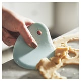 A hand uses a light blue LÄTTBAKAD spatula with a curved edge to scrape dough from a white surface.