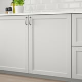 Light grey kitchen cabinets with beveled edges and handles, fitted to wooden floor and white tiled backsplash.