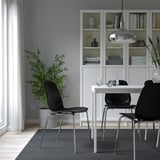 Modern dining setup with LIDÅS black chairs, white table, and pendant lamp. Grey rug on light wood floors. Large bookshelf with glass doors.