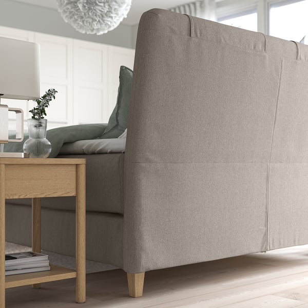 Modern grey upholstered bed with sloped headboard. Next to wooden table with vase and magazines. Bright, minimalist room.