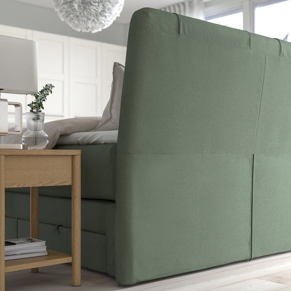 Green divan bed with cushioned headboard, featuring storage drawers below, set in a minimalist room with wooden floor and large window.