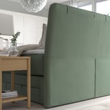 Green divan bed with cushioned headboard, featuring storage drawers below, set in a minimalist room with wooden floor and large window.