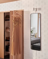LINDBYN mirror on white wall reflects bedroom with bed & closet of beige/black clothes, flowered dress.