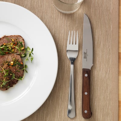 A dinner setting with a plate of steak, accompanied by a serrated steak knife and fork on a placemat.
