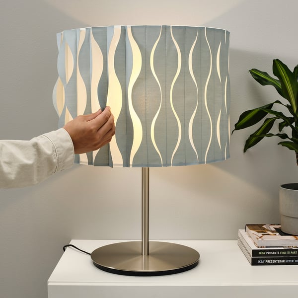 Person adjusting folded lampshade on modern brass base. The lamp sits on a white table with books and a plant nearby.