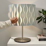 Person adjusting folded lampshade on modern brass base. The lamp sits on a white table with books and a plant nearby.