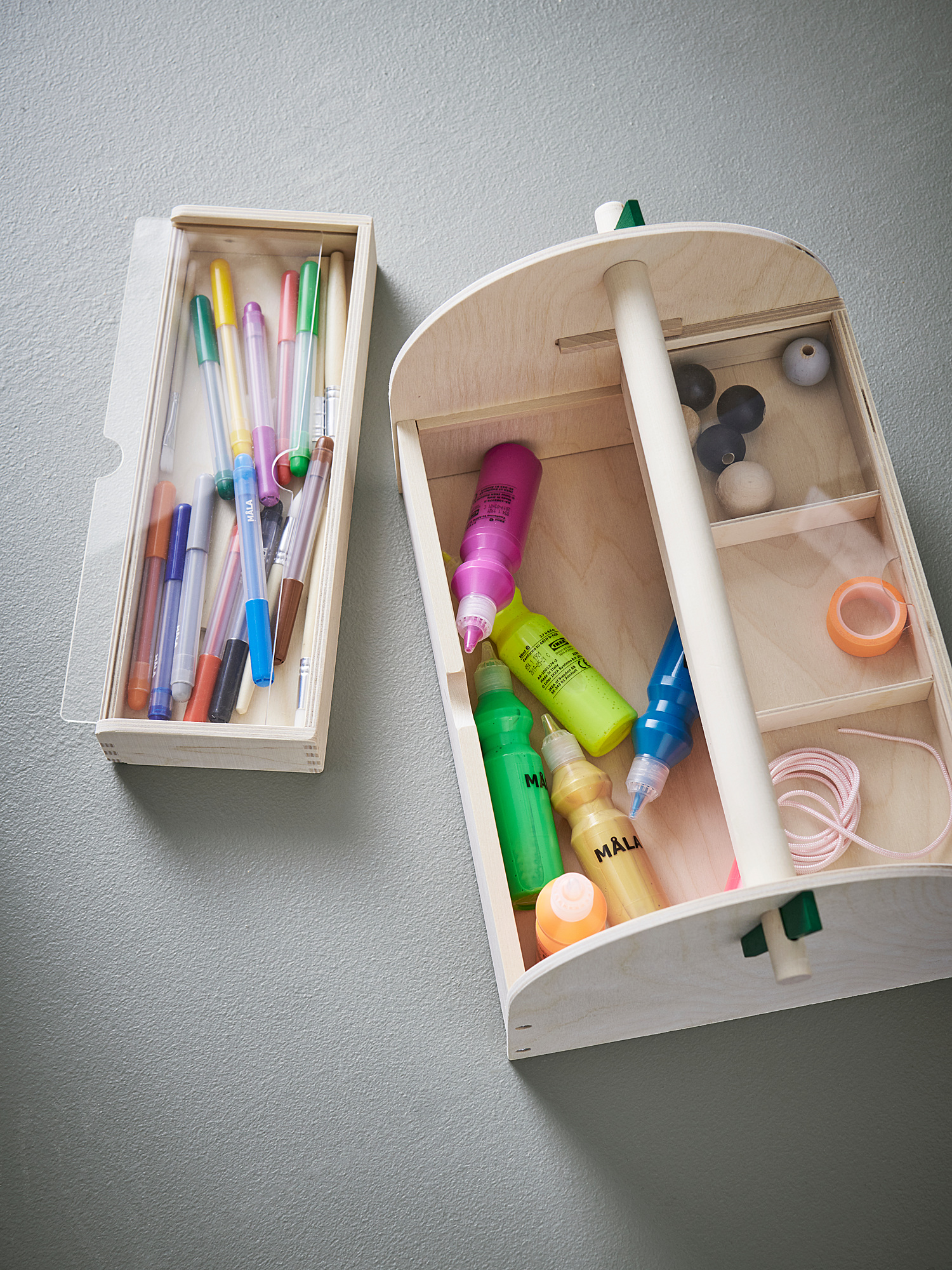 Two wooden storage boxes against a wall. One contains colorful pens. The other has art supplies, including paints, brushes, and tape.