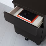Black MALM nightstand with open drawer showing notebook and pens, castors for easy movement.