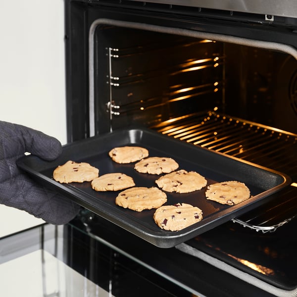 A person is placing a tray of chocolate chip cookies into an oven. The non-stick baking tray ensures easy release of baked goods.