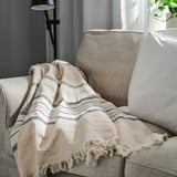 Beige fringe throw on grey couch, stripes, warm setting.