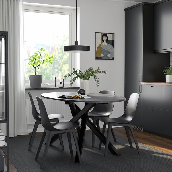 Modern dining setup with round table, four black chairs, pendant light, gray rug, and large window with natural light and artwork.