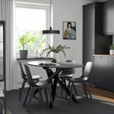 Modern dining setup with round table, four black chairs, pendant light, gray rug, and large window with natural light and artwork.