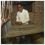 Man weaving striped bedspread on loom in rustic room.