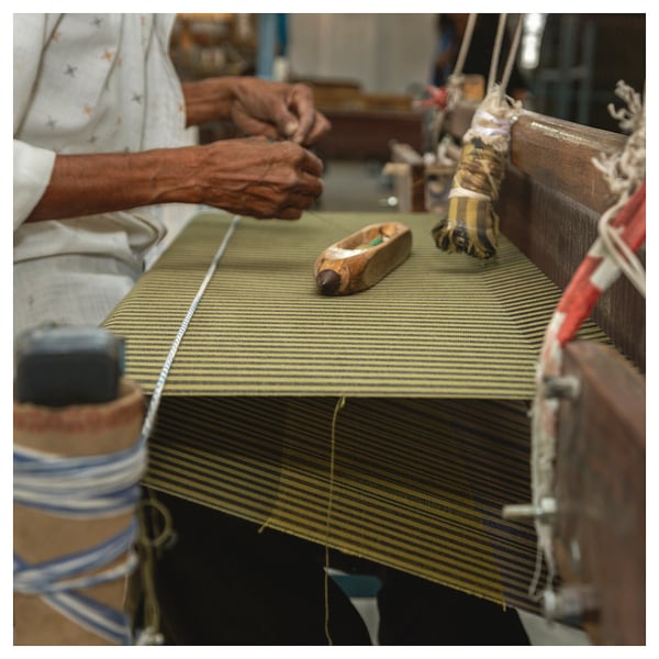 Person weaves yellow-striped MÄVINN bedspread on wooden loom. Vibrant stripes, cotton material, manual craftsmanship evident.