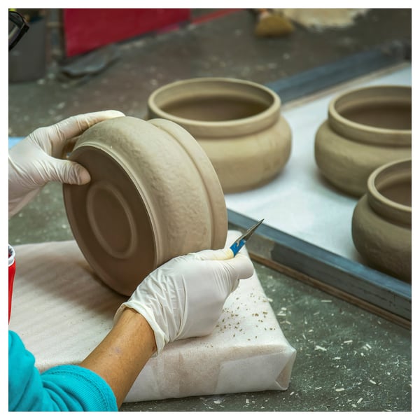 A craftsperson shapes clay bowls by hand, wearing gloves, with a tool. Several half-finished bowls rest nearby on a table.