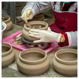 Person crafting pottery, wearing gloves and apron, using tools on clay bowl. Multiple bowls nearby on work surface.